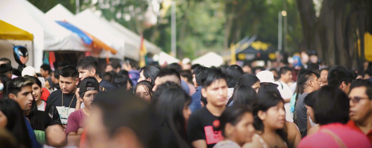 Na imagem consta vários grupos de pessoas conversando entre si. Ao fundo, há várias barracas dispostas uma ao lado da outra.