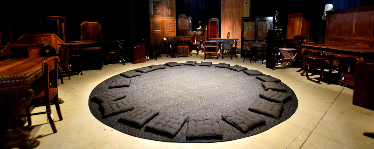 Na imagem constam almofadas pretas dispostas em círculo sobre tapete escuro, cercadas por móveis antigos de madeira.